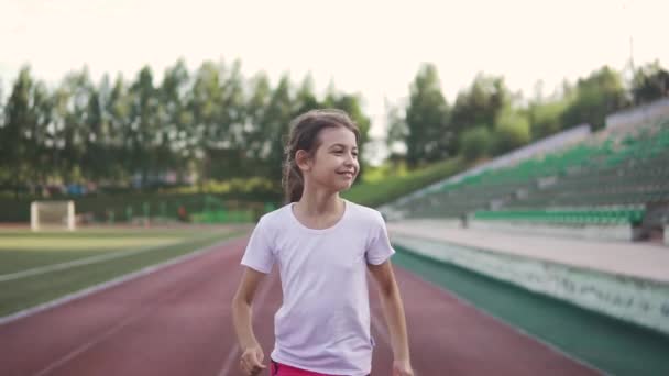 小さな女の子の肖像画は、ジョギングを楽しんでいます。子供が競技場でトラックに沿って走る - 映像、動画