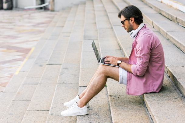 side view of man in sunglasses with headphones on neck using laptop while sitting on steps on street - Photo, Image