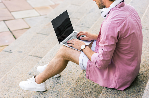 cropped shot of man with headphones on neck using laptop while sitting on steps on street - Photo, Image