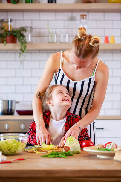 Kuva nuoresta äidistä tyttärensä kanssa leikkaamassa vihanneksia keittiössä
 - Valokuva, kuva