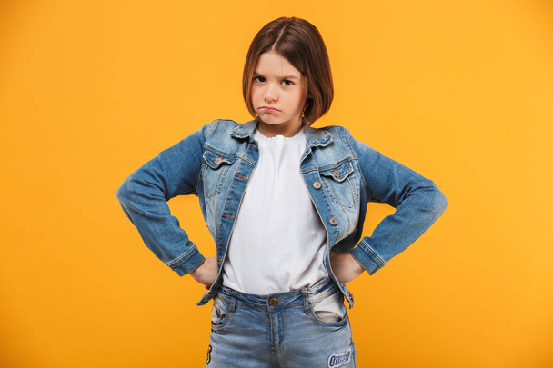 Portrait of an upset little schoolgirl standing with hands on hips over yellow background - Photo, image