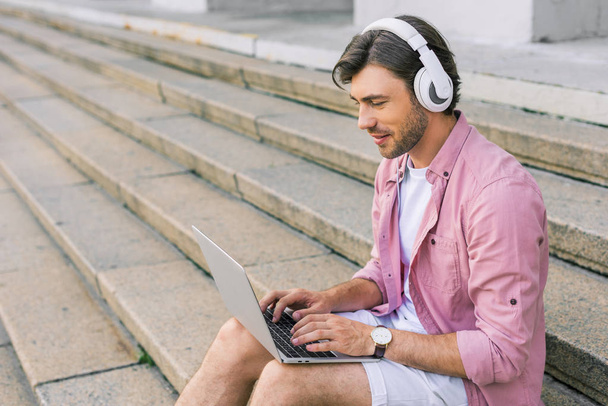 vista lateral del hombre con estilo en los auriculares usando el ordenador portátil mientras está sentado en los pasos en la calle
 - Foto, imagen