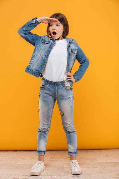 Full length portrait of an excited little schoolgirl standing and looking far away over yellow background - Foto, Imagen