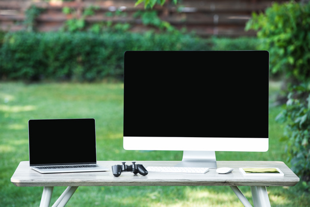 foco seletivo de joystick, tablet digital, computador e laptop com telas em branco na mesa ao ar livre
  - Foto, Imagem