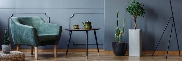 Small table with jug and tea cup standing next to green armchair in dark grey living room interior with molding on the wall and fresh green plants on the floor - Fotó, kép