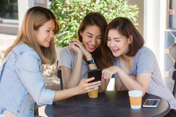 Веселі азіатські молоді жінки сидять в кафе п'ють каву з друзями і спілкуються разом. Приваблива азіатська жінка насолоджується кавою, використовуючи смартфон для розмов, читання та смс
. - Фото, зображення
