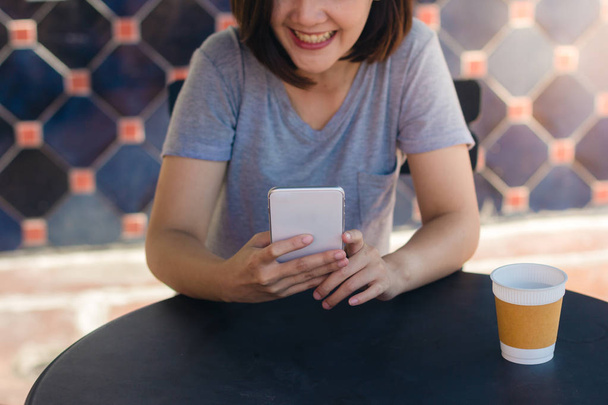 Joyeux asiatique jeune femme assise dans un café à boire du café et en utilisant un smartphone pour parler, lire et envoyer des SMS. Attrayant asiatique femme tenant une tasse de café tout en regardant sur son téléphone
. - Photo, image