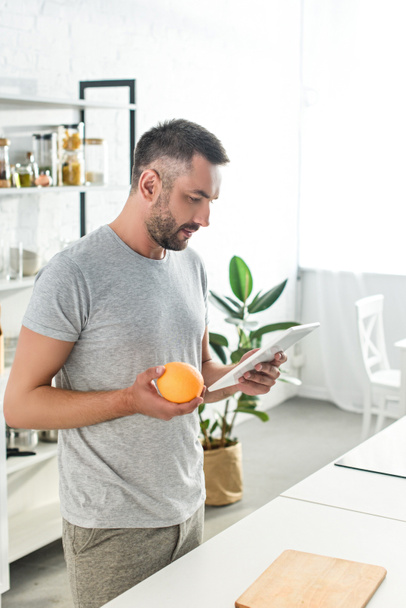 side view of man holding orange and looking for recipe in digital tablet on kitchen  - Fotografie, Obrázek