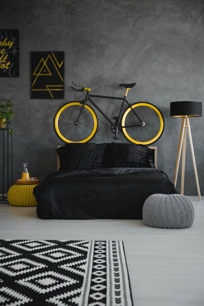 Bike above black bed in modern grey bedroom interior with patterned carpet, pouf and lamp. Real photo - Foto, Imagen