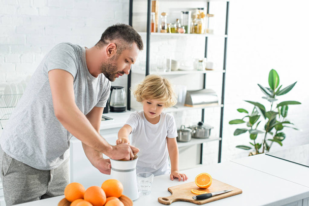 father and little son making fresh orange juice by squeezer on table at kitchen son  - Фото, изображение