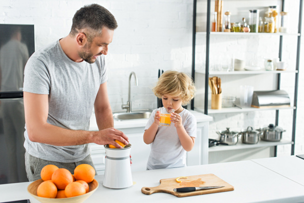padre haciendo jugo de naranja por exprimidor mientras su pequeño hijo bebiendo jugo en la cocina
  - Foto, imagen