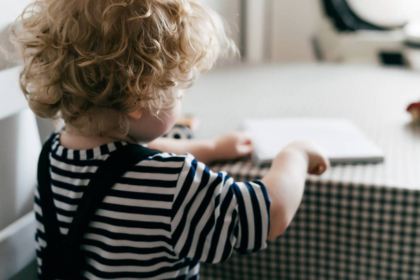curly blond little boy in cute clothes sitting at a table and drawing in a notebook - Photo, Image