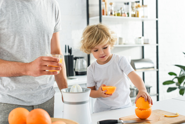 フレッシュ ジュースと台所でまな板からオレンジを取って彼の息子のガラスとの父親のイメージをトリミング - 写真・画像