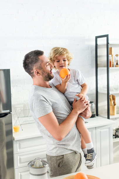 キッチンで手でオレンジ色の小さな息子を保持している大人の男 - 写真・画像
