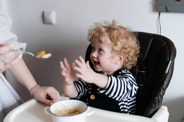 caring mother feeds her little curly son with a useful hot soup in the kitchen - Zdjęcie, obraz
