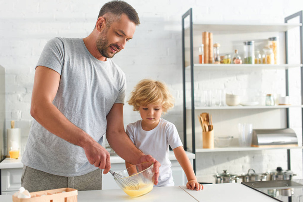 усміхнений батько збиває яйця в мисці, а його син стоїть біля кухні
  - Фото, зображення