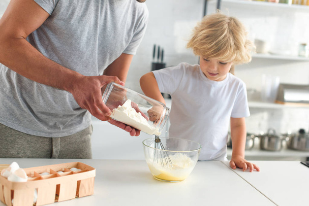 cropped image of man pouring flour into bowl while his son whisking eggs at kitchen  - 写真・画像