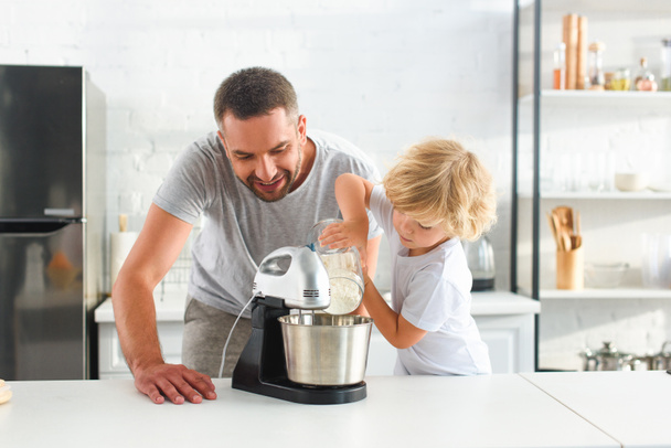 маленький хлопчик поливає борошно в миску мікшера, а його батько стоїть біля кухні
  - Фото, зображення