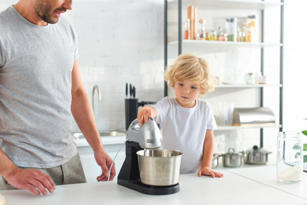 περικομμένη εικόνα του ανθρώπου που στέκεται κοντά γιος ενώ ο ίδιος χρησιμοποιώντας το μίξερ στην κουζίνα  - Φωτογραφία, εικόνα