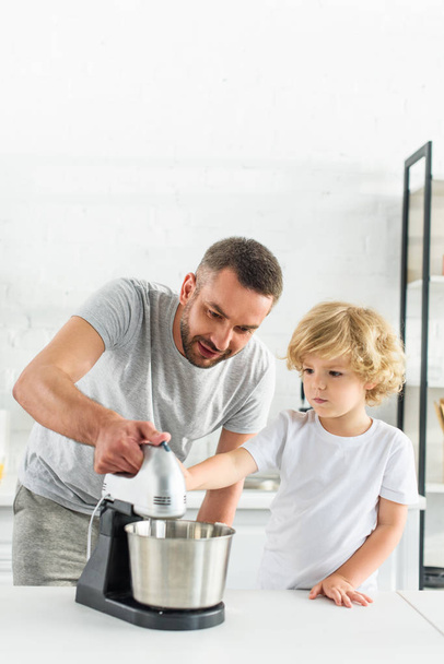father teaching son using mixer on tabletop at kitchen - Photo, Image