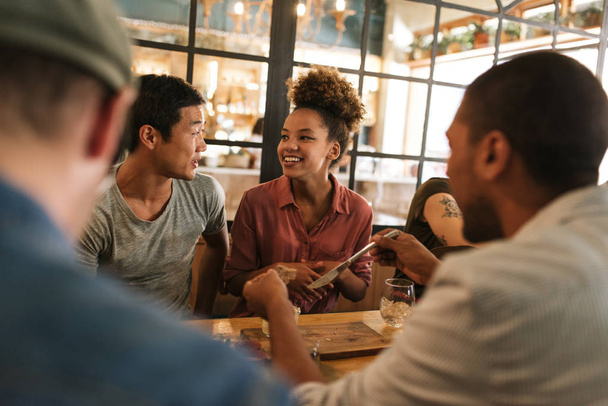 Zróżnicowaną grupę uśmiechający się młodzi przyjaciele siedzi przy stole w bistro, rozmawiając ze sobą ponad posiłek - Zdjęcie, obraz
