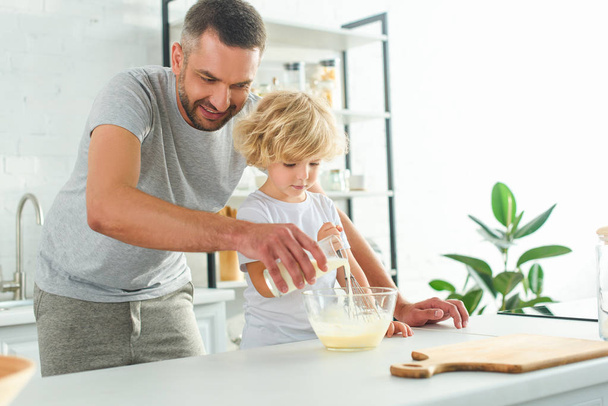 adorable boy whisking dough while his father pouring milk into bowl at kitchen - Foto, Imagen