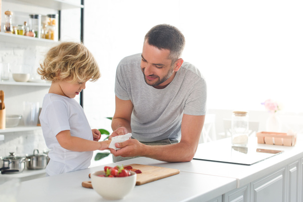 キッチンでイチゴと卓上近くナプキンで拭く手に息子を助けて笑顔の父  - 写真・画像