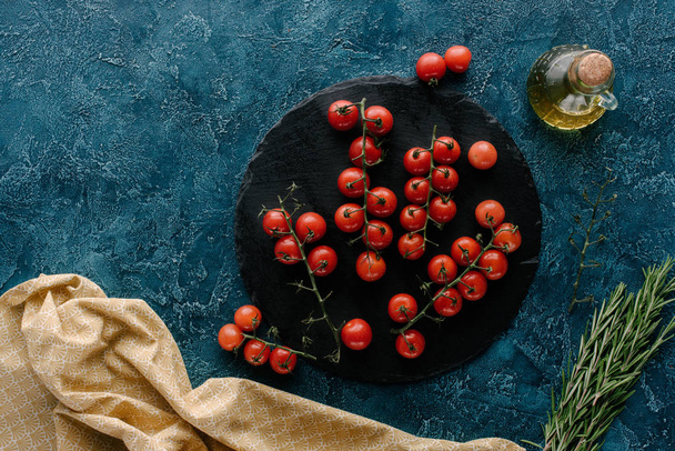 Placa de ardósia escura com tomate cereja e garrafa de óleo na mesa azul
 - Foto, Imagem