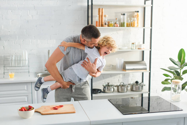 father holding laughing son and tickling him at kitchen - Photo, Image