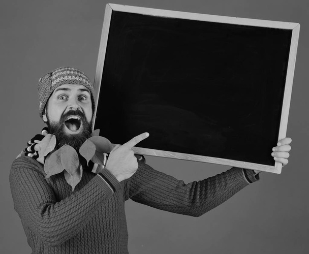 Guy points at chalkboard at barbershop, copy space. Hipster with excited face and beard full of leaves. Autumn fashion ad and presentation concept. Man in warm hat - Fotografie, Obrázek