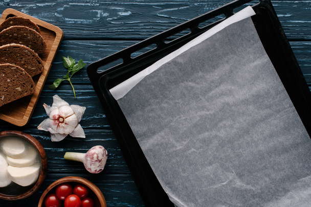 Empty baking sheet on dark wooden table by cooking ingredients and tomatoes - Фото, зображення