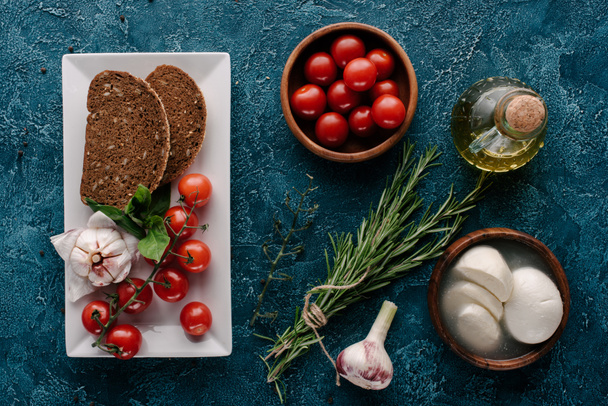 Mozzarella cheese and tomatoes on dark blue table with bread and herbs - Фото, зображення