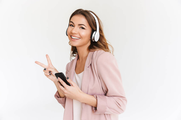 Image of young cute beautiful woman isolated over white wall background listening music with headphones using mobile phone make peace gesture. - Valokuva, kuva