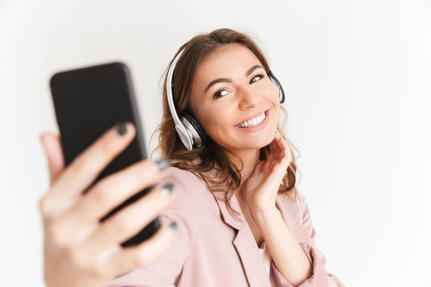 Image of young cute beautiful woman isolated over white wall background listening music with headphones make selfie by mobile phone. - Фото, изображение