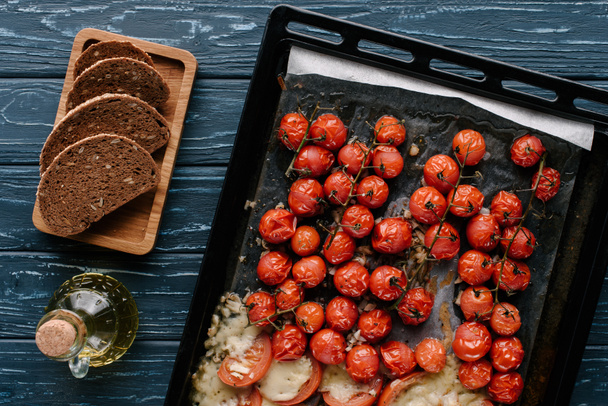 Tomates cereja vermelhos assados com queijo em mesa de madeira escura com pão e óleo
 - Foto, Imagem