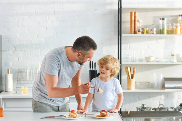 padre sonriente alimentando al pequeño hijo con panqueques en la cocina
 - Foto, imagen