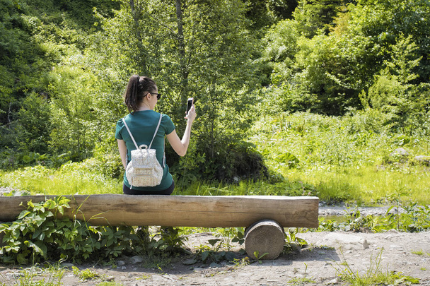 Menina com uma mochila perto da floresta tira uma foto em um smartphone. Vista traseira. Verão dia ensolarado
 - Foto, Imagem