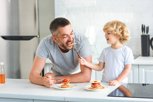 Улыбающийся маленький мальчик кормит отца блинами на кухне
 - Фото, изображение