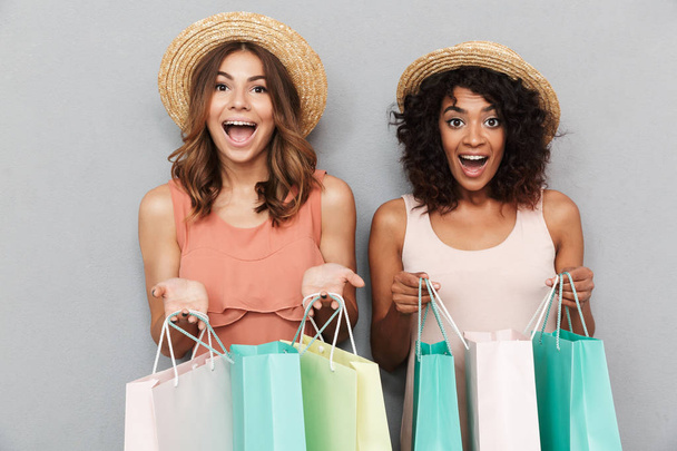 Portrait of two happy young women dressed in summer clothes holding shopping bags isolated over gray background - Foto, Imagem