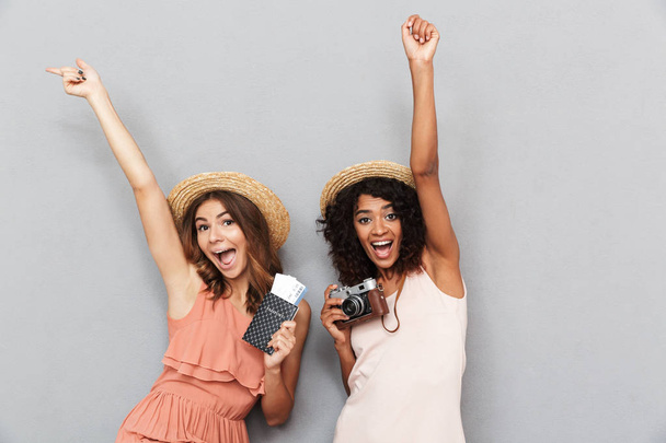 Portrait of two happy young women dressed in summer clothes holding passport with tickets and celebrating over gray background - Foto, Bild