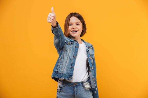 Portrait of a cheerful little schoolgirl showing thumbs up gesture over yellow background - Photo, image