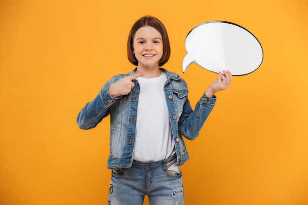 Portrait of a happy little schoolgirl holding empty speech bubble and pointing finger over yellow background - Photo, Image