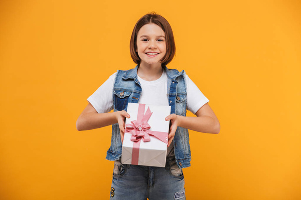 Portrait of a happy little schoolgirl holding gift box over yellow background - Фото, изображение