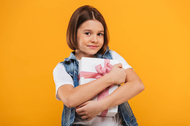 Portrait of a satisfied little schoolgirl hugging gift box over yellow background - Φωτογραφία, εικόνα