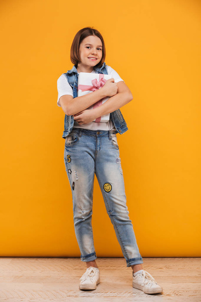 Full length portrait of a satisfied little schoolgirl holding gift box over yellow background - Foto, Bild