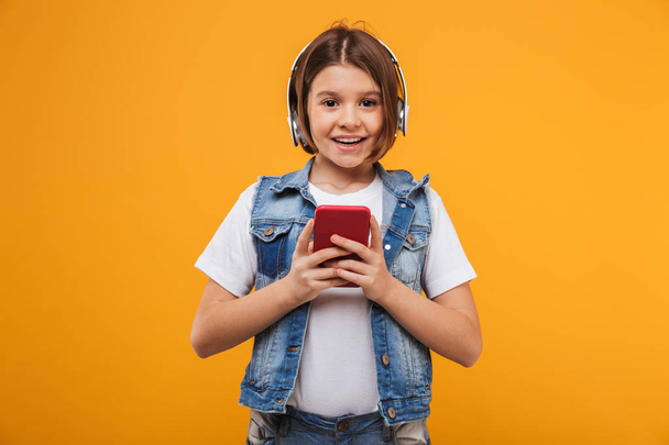 Portrait of a joyful little schoolgirl listening to music with headphones while holding mobile phone over yellow background - Foto, Bild