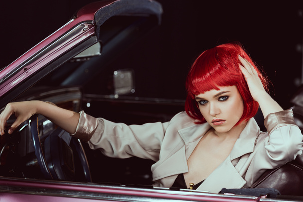 όμορφη νεαρή γυναίκα σε κόκκινη περούκα και κομψή καμπαρντίνα κάθεται στο αυτοκίνητο και να βλέπουν τα φωτογραφικών μηχανών - Φωτογραφία, εικόνα
