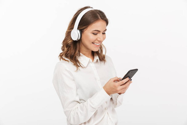 Portrait of a smiling young business woman listening to music with headphones while using mobile phone isolated over white background - Fotoğraf, Görsel