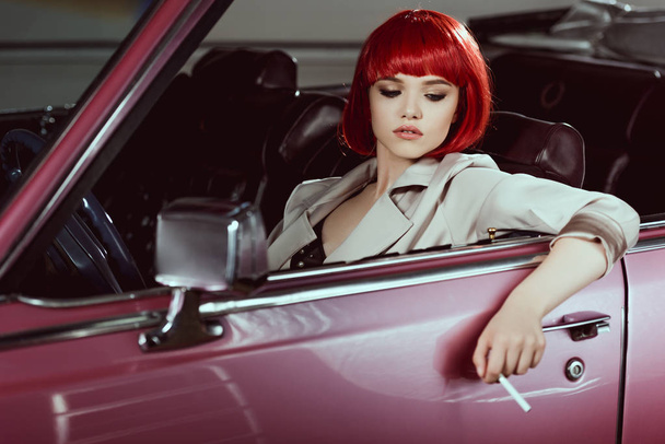 beautiful stylish girl in red wig holding cigarette and sitting in vintage car  - 写真・画像