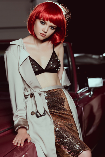 beautiful young woman in red wig, bra and trench coat sitting on classic car and looking at camera - Foto, Bild